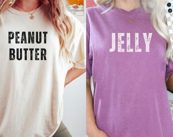 May include: Two women wearing matching t-shirts. One shirt is cream colored with the text "PEANUT BUTTER" in black. The other shirt is purple with the text "JELLY" in white.