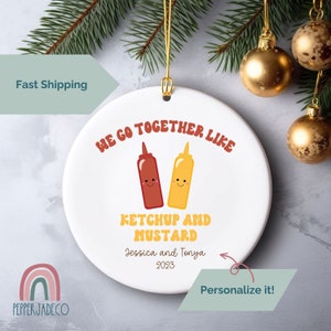 May include: White ceramic ornament with a red and yellow cartoon ketchup and mustard bottle design. The text reads "We go together like ketchup and mustard" with the option to personalize with names and a year.