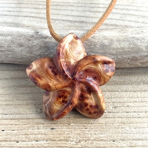 May include: A wooden pendant in the shape of a flower with brown and gold swirls. The pendant is on a brown suede cord.