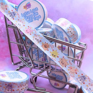 May include: A collection of light blue washi tape rolls with cartoon animal designs and the text "Lotsa Love Skzoo 5m Washi Tape." The tape features illustrations of a rabbit, bear, fox, and other characters. The rolls are in a miniature shopping cart.