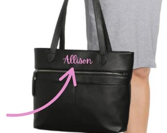 May include: Black tote bag with two zippered pockets and long handles. The name "Allison" is written in pink script on the front of the bag. The bag is held by a person wearing a gray dress and black heels.
