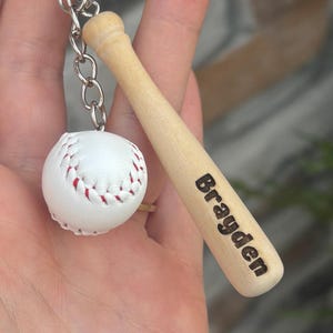 May include: A baseball-themed keychain featuring a miniature white baseball with red stitching and a small wooden bat. The bat is engraved with the name "Brayden". The keychain is attached to a silver chain.
