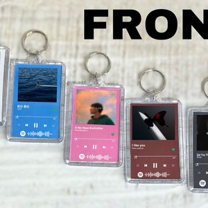 May include: Five clear acrylic keychains with different Spotify song covers. The keychains feature a house with the text "Stuck With U", a blue ocean with the text "KII BII", a pink sunset with the text "If We Have Eachother", a dark background with a red butterfly and the text "I Like You", and a dark background with the text "Get You The Moon".