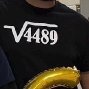 May include: Black crew neck t-shirt with the white text "√4489" printed on the front. The shirt is made of a soft material. A gold balloon is visible in the foreground.