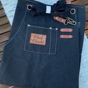 May include: Black denim apron with brown leather accents and the text "Chef Chadd" on a leather patch. Features a pocket, adjustable neck strap, and a metal clasp. The apron is designed for kitchen use.