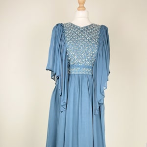 May include: A long, flowing blue dress with a beaded bodice and sheer sleeves. The dress is displayed on a mannequin, highlighting its elegant design and draping fabric. The bodice features intricate beadwork.