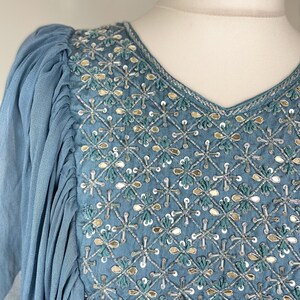 May include: A pale blue dress with a V-neck and detailed embroidery. The bodice has a diamond pattern with silver, gold, and teal embellishments. The sleeves are gathered at the shoulder and flow down the arm.