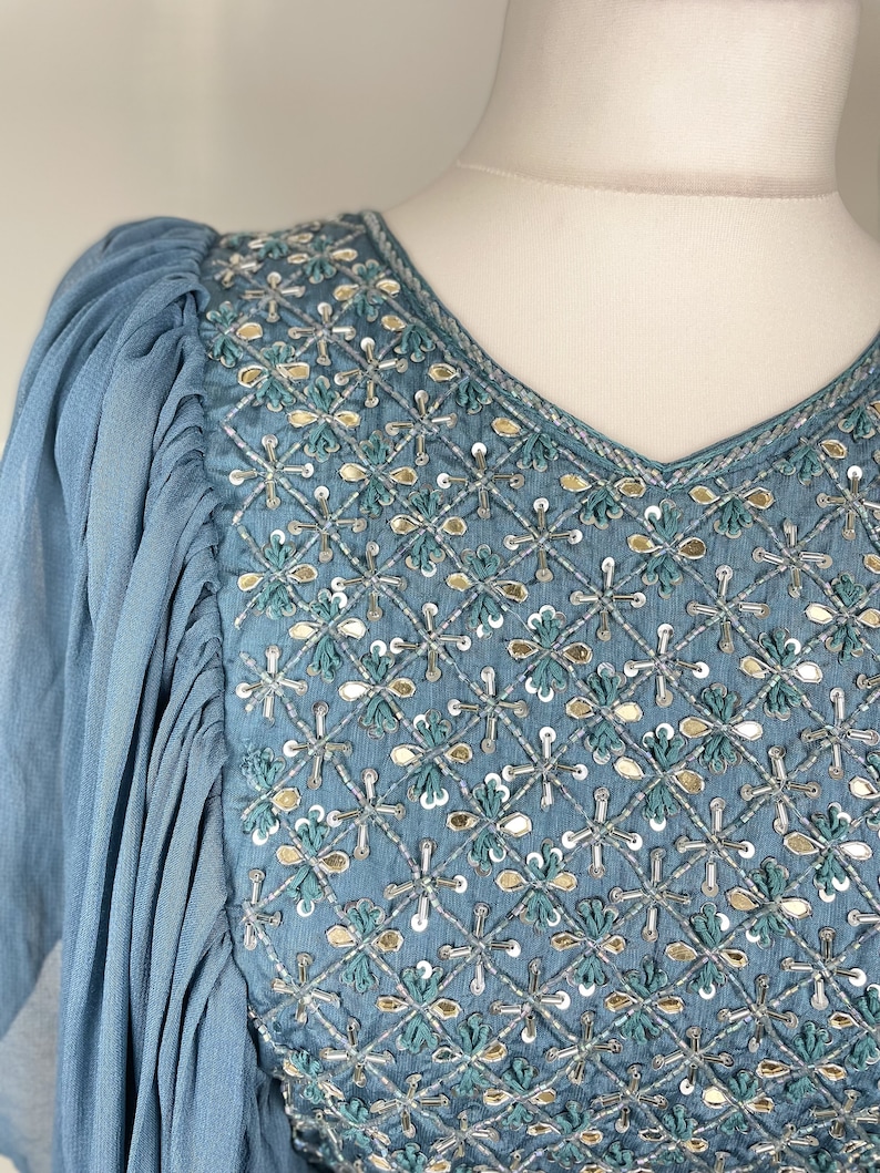 May include: A pale blue dress with a V-neck and detailed embroidery. The bodice has a diamond pattern with silver, gold, and teal embellishments. The sleeves are gathered at the shoulder and flow down the arm.