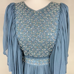May include: A light blue dress with a V-neck and long, flowing sleeves. The bodice is adorned with detailed embroidery and sequins in a floral design. The dress has a cinched waist and a flowing skirt, creating an elegant silhouette.