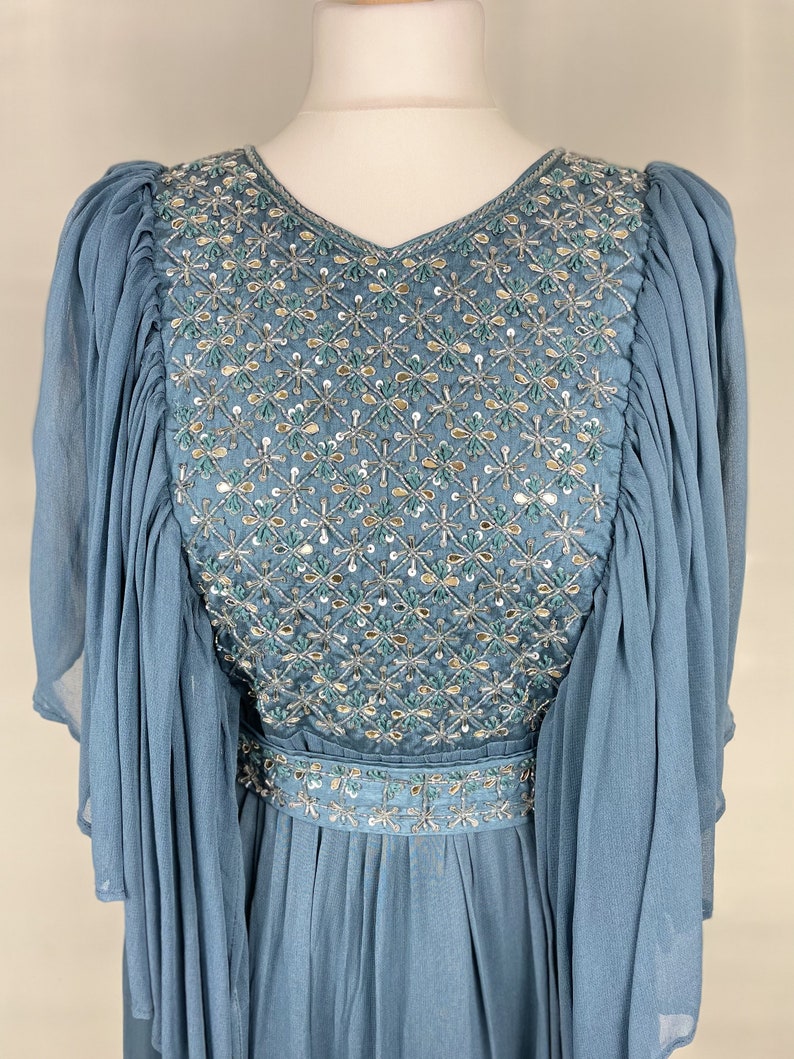 May include: A light blue dress with a V-neck and long, flowing sleeves. The bodice is adorned with detailed embroidery and sequins in a floral design. The dress has a cinched waist and a flowing skirt, creating an elegant silhouette.