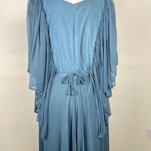 May include: A powder blue dress with a flowing design. The back of the dress has a zip and a tie at the waist. The sleeves are made of a sheer, layered fabric, adding a touch of elegance. The dress is displayed on a mannequin.