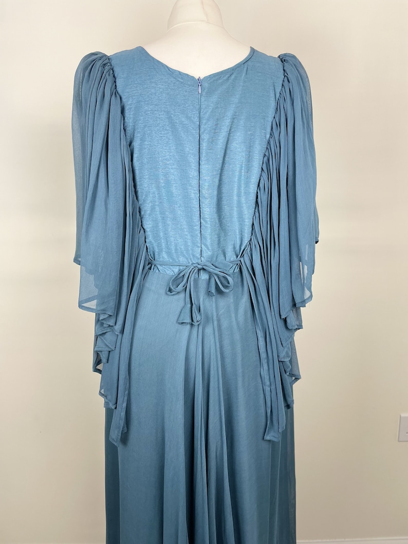 May include: A powder blue dress with a flowing design. The back of the dress has a zip and a tie at the waist. The sleeves are made of a sheer, layered fabric, adding a touch of elegance. The dress is displayed on a mannequin.
