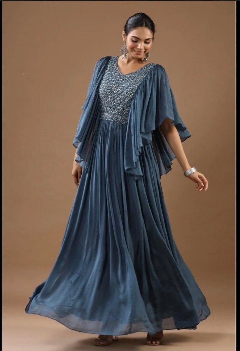 May include: A long, slate blue gown with a V-neck and matching cape. The bodice is adorned with silver sequins. The dress features a layered, pleated skirt and flared sleeves. The model is wearing silver earrings and a bracelet. The dress is floor-length.
