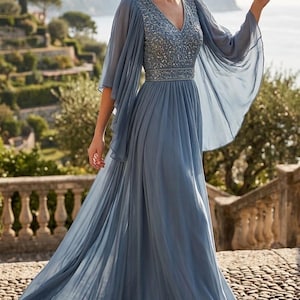 May include: A long, dusty blue formal dress with a V-neck and embellished bodice. The dress has long, flowing sleeves and a full skirt. The model is standing on a stone path with a scenic background.