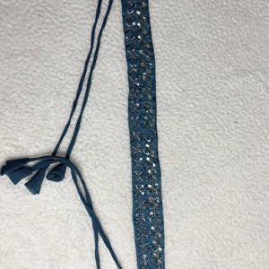 May include: Teal headband featuring silver sequin floral detailing. The headband has long, teal ties at each end, finished with small tassels. The decorative strip is approximately 2.5 cm wide.