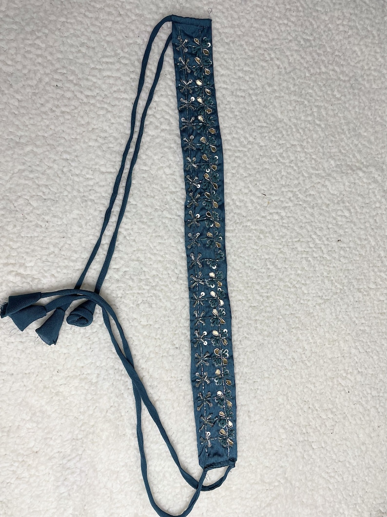 May include: Teal headband featuring silver sequin floral detailing. The headband has long, teal ties at each end, finished with small tassels. The decorative strip is approximately 2.5 cm wide.