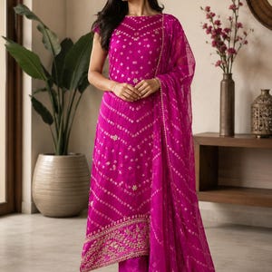 Puede incluir: Un conjunto de kurta sin mangas en color magenta vibrante con un patrón de tie-dye bandhani blanco en toda la tela. El dobladillo de la kurta está adornado con intrincados bordados dorados. Un dupatta magenta transparente a juego con borde dorado cuelga sobre un hombro.