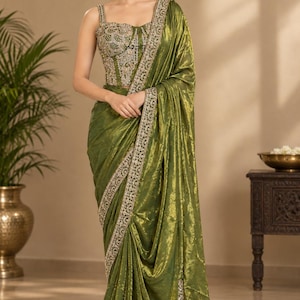May include: An olive green saree featuring a shimmering, textured fabric with a draped silhouette. The bodice is a structured, sleeveless corset-style top adorned with intricate silver embroidery, sequins, and beadwork along the neckline and hem.