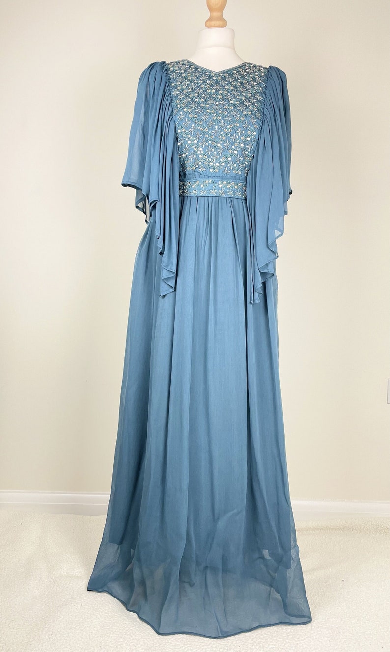 May include: A long, flowing blue dress with a beaded bodice and sheer sleeves. The dress is displayed on a mannequin, highlighting its elegant design and draping fabric. The bodice features intricate beadwork.