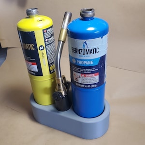 May include: A gray metal stand holds two propane tanks, one yellow and one blue. The yellow tank is labeled "MAP/PRO" and the blue tank is labeled "BERNZOMATIC" with the text "PROPANE" and "SINCE 1876".