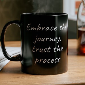 May include: Black ceramic coffee mug with the text "Embrace the journey, trust the process" printed in white on the side. The mug is sitting on a wooden table with a newspaper and a glass teapot in the background.