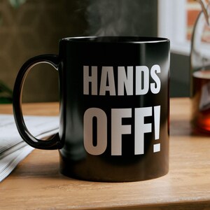 May include: Black ceramic mug with white text that reads "HANDS OFF!" The mug is filled with a hot beverage and steam is rising from the top.