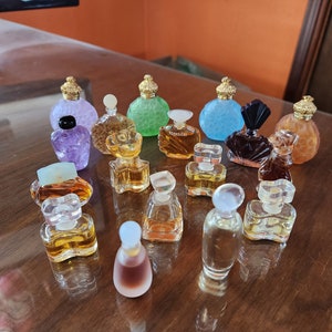Pode incluir: Uma coleção de frascos de perfume em miniatura de várias cores e formas, incluindo transparente, verde, azul, roxo e marrom. Os frascos estão dispostos sobre uma superfície de madeira.