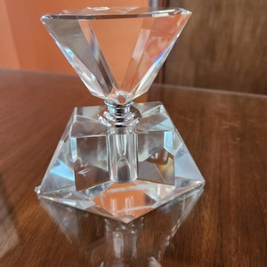 Peut inclure: Un flacon en verre transparent avec un bouchon en forme de diamant facetté et un capuchon en métal argenté. Le flacon est posé sur une surface en bois.