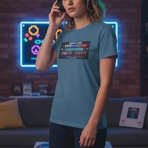 May include: A slate blue short-sleeved t-shirt featuring a rectangular graphic print on the chest. The graphic displays a series of directional arrows and buttons above the text "KONAMI CODE" and "CHEATERS GONNA CHEAT" in a neon-style font.