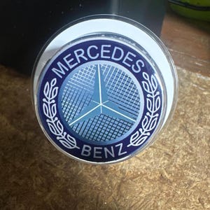 May include: A round emblem featuring the Mercedes-Benz logo. The logo includes a three-pointed star within a circle, surrounded by the words "MERCEDES" and "BENZ" in white text on a dark blue background. The emblem is framed by a white border.