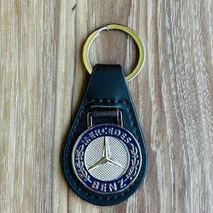 Op de afbeelding: Een zwarte leren sleutelhanger met een zilveren Mercedes-Benz-logo. Het logo is omringd door een blauwe en zilveren krans.