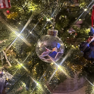 May include: A clear glass Christmas bauble filled with small, colourful folded paper decorations. The bauble is hanging on a green Christmas tree with white fairy lights.
