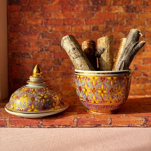 May include: A decorative ceramic jar with a lid, featuring a vibrant floral pattern in yellow, pink, and gold. The jar is filled with several pieces of wood. The lid is placed next to the jar.