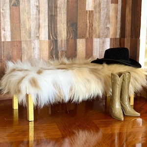 May include: A miniature bench with a faux fur top in shades of cream and tan, supported by gold-colored legs. A black cowboy hat and a pair of tan cowboy boots are placed on the bench, creating a Western-themed display.