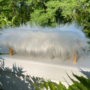May include: A white faux fur bench with wooden legs. The bench is sitting on a white surface.