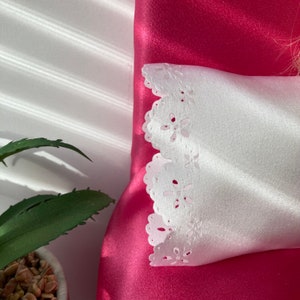 May include: A white satin pillow with a delicate lace trim. The pillow is on a bright pink satin surface.