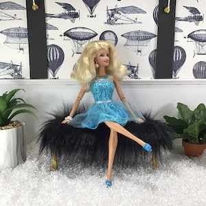 May include: A doll wearing a blue lace dress and blue shoes sits on a black furry bench. The doll has blonde hair and blue eyes. The bench is on a white surface with a white and black patterned background.