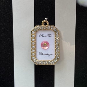 Puede incluir: Un amuleto rectangular dorado con fondo blanco y un botón rosa en el centro. El amuleto está rodeado de diamantes de imitación transparentes y perlas blancas. El texto "Press For Champagne" está impreso en el amuleto.