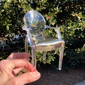 May include: A clear plastic chair with a gold-colored seat and a gold letter "Q" on the back. The chair is being held in a hand.