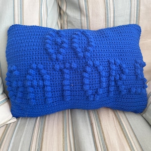 Puede incluir: Una almohada de punto de crochet azul con un diseño texturizado y abultado. La almohada es rectangular y tiene un aspecto suave y acogedor.