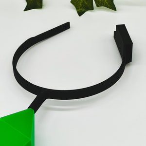 May include: A black headband with a green diamond-shaped pendant. The headband is made of a flexible material and is designed to fit comfortably on the head.