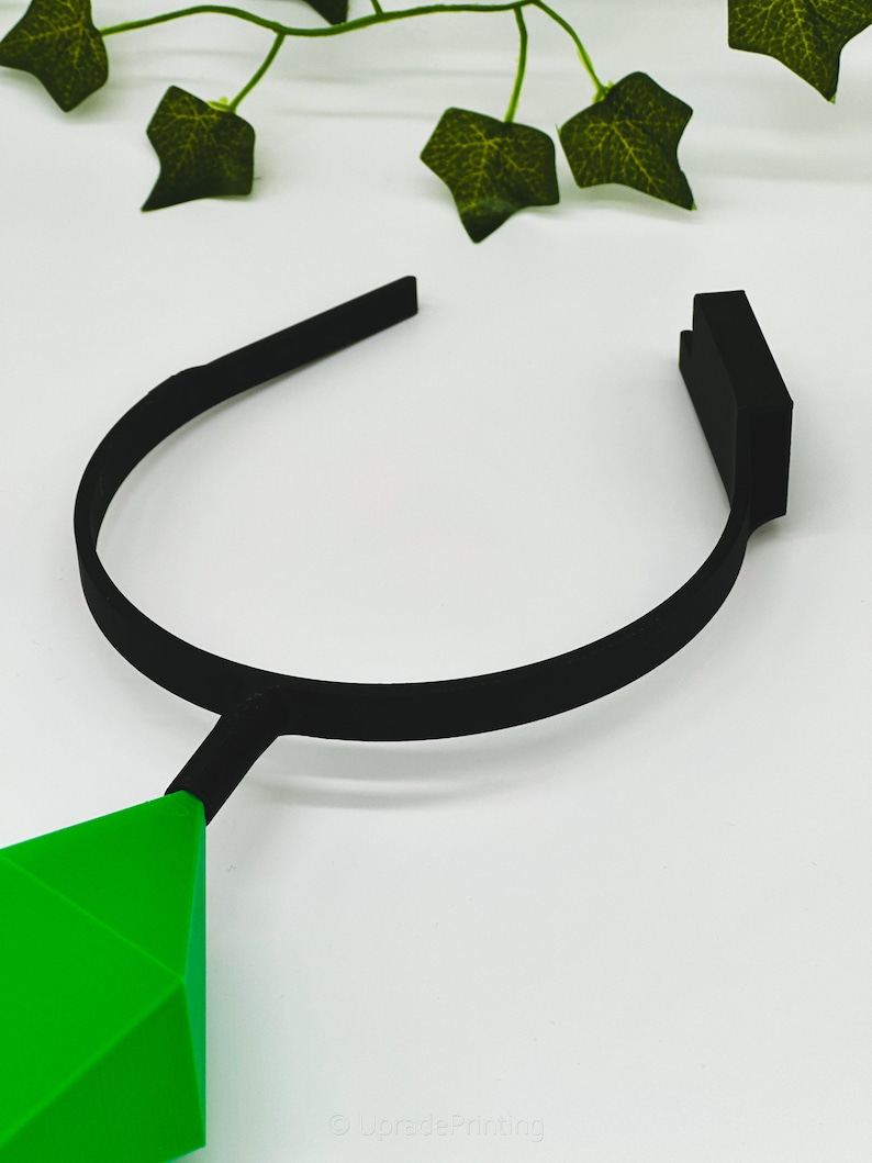May include: A black headband with a green diamond-shaped pendant. The headband is made of a flexible material and is designed to fit comfortably on the head.