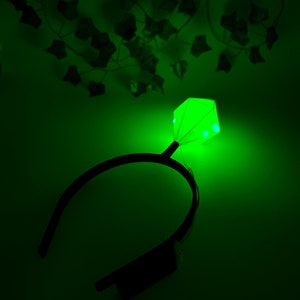 May include: A green light-up headband with a diamond-shaped light in the center. The headband is black and the light is a bright green.