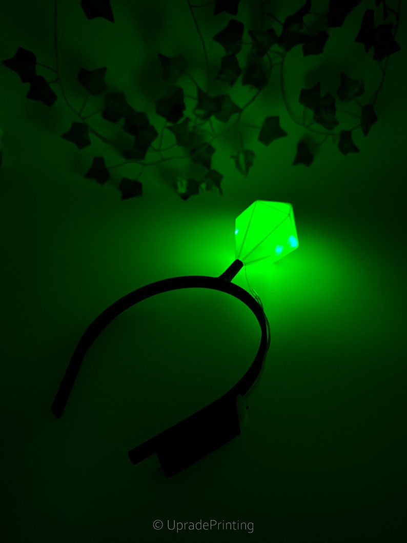 May include: A green light-up headband with a diamond-shaped light in the center. The headband is black and the light is a bright green.