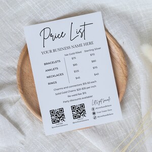 May include: A white price list on a wooden plate, featuring the text "Price List" and "YOUR BUSINESS NAME HERE." It lists prices for bracelets, anklets, necklaces, and rings in both 14k gold-filled and sterling silver. Includes QR codes and social media handles.