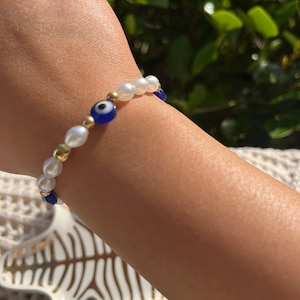 May include: A bracelet with white pearls, gold beads, and a blue evil eye charm. The bracelet is strung on a thin cord and is designed to be worn on the wrist. The bracelet is a stylish accessory, perfect for everyday wear.