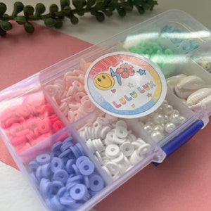 May include: A clear plastic organizer filled with colorful polymer clay beads, pearls, and seashells. The beads are in shades of pink, white, blue, and green. A sticker with a smiling face and the text "Lulu Lux" is on the lid.