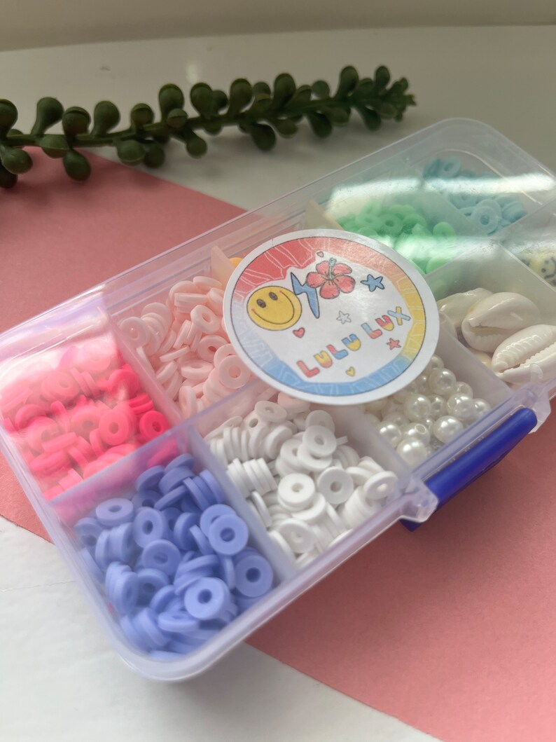 May include: A clear plastic organizer filled with colorful polymer clay beads, pearls, and seashells. The beads are in shades of pink, white, blue, and green. A sticker with a smiling face and the text "Lulu Lux" is on the lid.