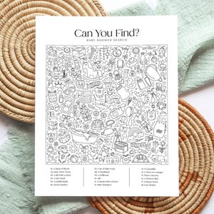 May include: Black and white baby shower search game with the text "Can You Find?" and a list of items to find. The game is surrounded by woven placemats and a green cloth.