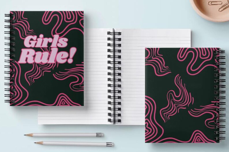 May include: Two spiral bound notebooks with black covers and pink abstract line designs. One notebook has the text "Girls Rule!" in pink letters.